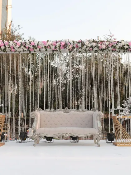 A chic and modern stage for a day wedding, with a backdrop of shimmering silver strings and a lush floral border. The classic white sofa completes the look.