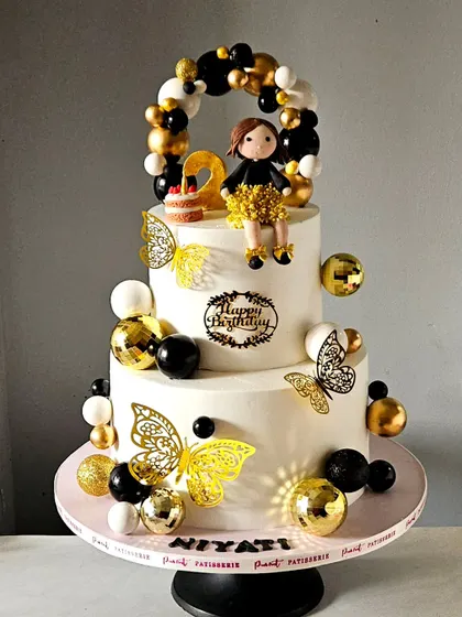 A chic and glamorous cake in a black, gold, and white theme for a second birthday. The design features a balloon garland, shimmering butterflies, and a custom-made girl figurine in a sparkling dress.