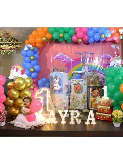 A fun Cocomelon and Unicorn combination theme for a first birthday party. This setup uses a rainbow of balloons, character-printed plinths, and a large light-up number '1'.