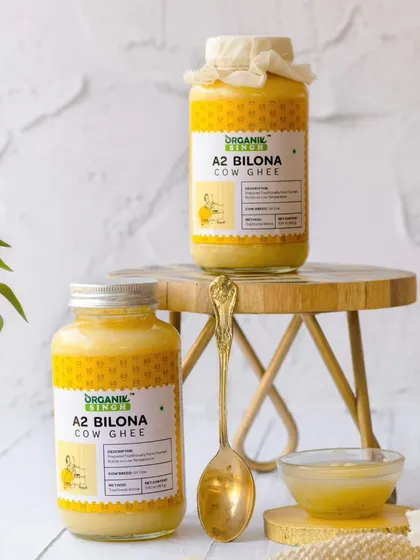 Two jars of A2 Bilona Cow Ghee are styled in a bright, airy setting on a small wooden stool. The clean background and touch of greenery give this shot a fresh, modern, and healthy feel.