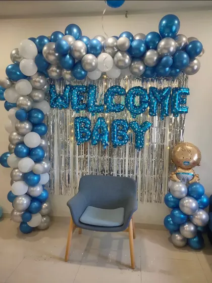 A simple and elegant 'Welcome Baby' setup with a blue, silver, and white balloon arch over a silver shimmer curtain.