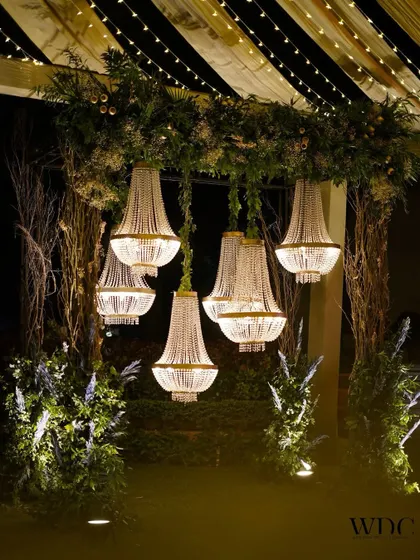 Shimmering chandeliers hanging amidst lush greenery and rustic branches, creating a beautiful contrast of glamour and nature for the cocktail night.