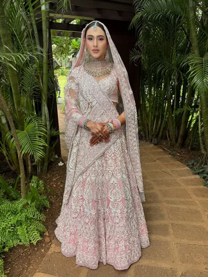 The full bridal look against a lush green backdrop. The intricate white embroidery on her pink lehenga is breathtaking, and the makeup was designed to enhance her natural beauty without distracting from the outfit's details.