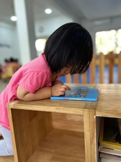 A child stands at a low bookshelf, pointing at pictures in a book. Our child-sized furniture makes all learning materials accessible.