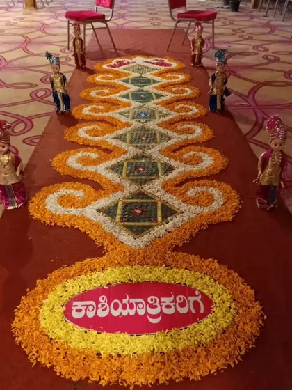 An identical image of the detailed floral rangoli for the Kashi Yatre ritual, flanked by traditionally dressed dolls, adding to the charm of the ceremony.