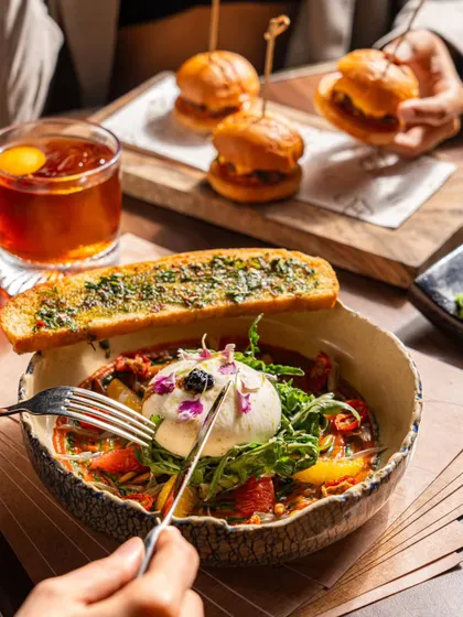 A lifestyle shot of a person cutting into a creamy burrata salad, with mini burgers and a cocktail nearby. This captures the interactive and social aspect of dining.