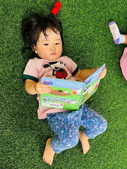 A toddler enjoys some quiet time, lying on the grass and looking through a Peppa Pig book. We encourage picture reading to build early literacy habits.