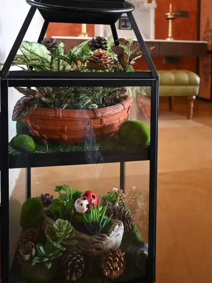The final result of our candle stand transformation. The bottom shelf features a miniature landscape with artificial succulents and moss, while the top holds a basket of live, low-light plants, creating a unique open terrarium.