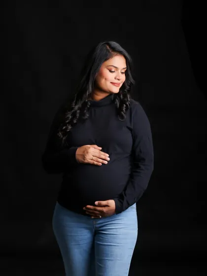 A simple and beautiful portrait of a woman in a black top and jeans, cradling her bump against a solid black background. Her gentle smile and the classic styling create a warm and timeless feel.