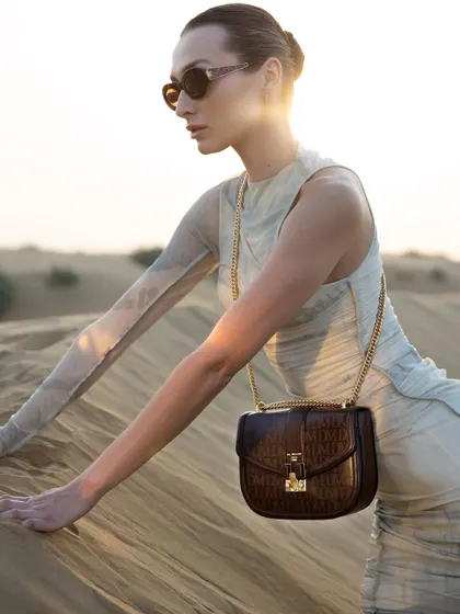 A sunset shot from the Da Milano desert campaign. The model is styled in a semi-sheer, ruched dress, with the warm light catching the texture of both the garment and the brown leather crossbody bag.