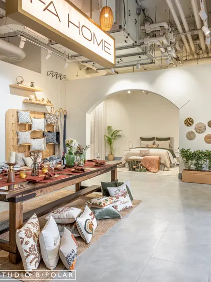 A beautifully set dining table serves as a central merchandising point, showcasing FA Home's table linens and decor in a real-life context. The floor is scattered with cushions, inviting customers to touch and feel the products.