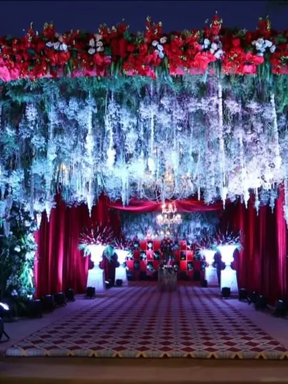 A grand and dramatic entrance for a reception. The walkway is defined by a ceiling of hanging white and blue flowers, framed by lush greenery and rich red draping for a luxurious feel.