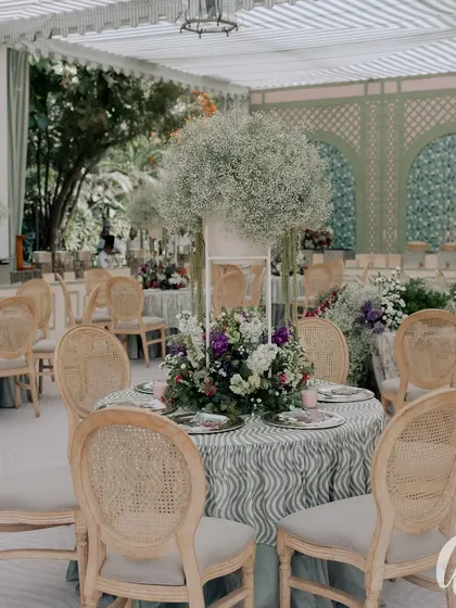 A beautifully set table featuring a towering baby's breath centerpiece. The patterned tablecloth and classic chairs create an elegant and timeless look for this garden event.