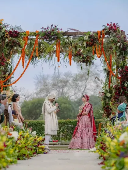 The couple exchanges vows under a lush floral mandap. The aisle is lined with wildflowers, and grand chandeliers hang overhead, crafting a perfect moment where love is sealed.
