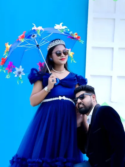A stylish couple's maternity portrait, with both wearing sunglasses and posing against a bright blue backdrop.