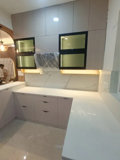 An in-progress shot from our luxury 3BHK project in ATS Rhapsody. This shows the L-shaped layout with light-colored Century HDHMR cabinets, a white quartz countertop, and a marble-patterned backsplash. The under-cabinet lighting is already installed to provide task lighting.