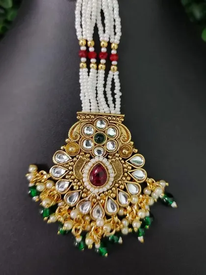 A traditional maang tikka with a central ruby stone, surrounded by kundan and green bead drops. The white pearl strands add to its classic appeal.