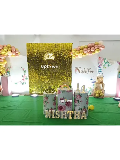 A birthday setup with a gold sequin backdrop and floral-themed pedestals.