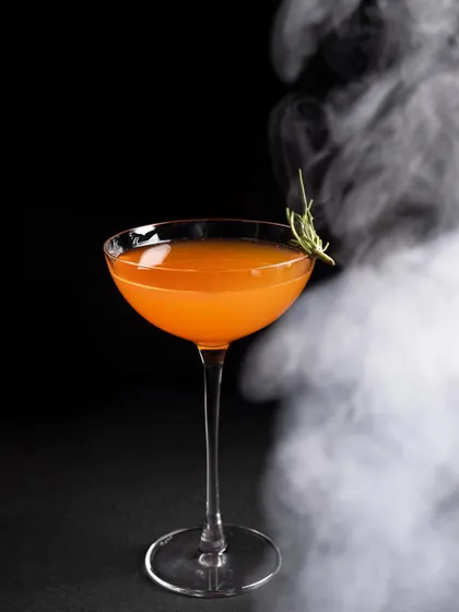 A single orange cocktail with a rosemary garnish, with a plume of smoke artfully swirling around it for a cinematic effect.