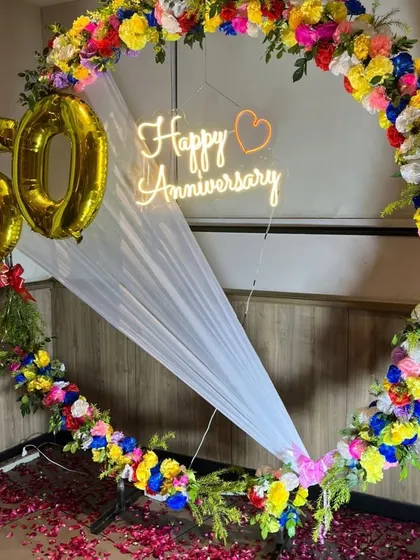A grand 50th-anniversary decoration featuring a large ring covered in colorful artificial flowers, a 'Happy Anniversary' neon sign, and '50' foil balloons.