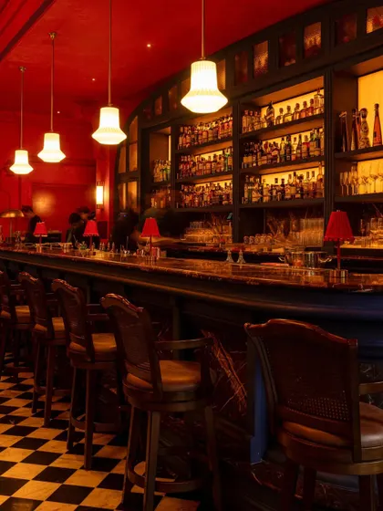 The main bar at BANNG, where a classic wooden structure is set against a vibrant red backdrop and a checkerboard floor. The design fuses traditional bar aesthetics with a bold, contemporary color narrative, creating a central point of energy in the restaurant.