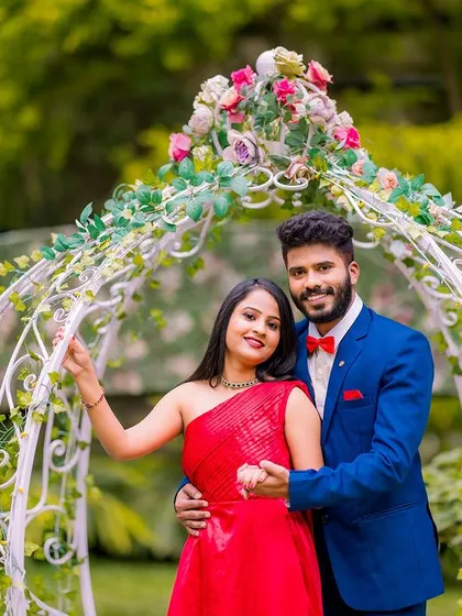 A happy couple poses in front of a flower-adorned arch and carriage. The bride's red dress and the groom's blue suit create a vibrant and stylish look.