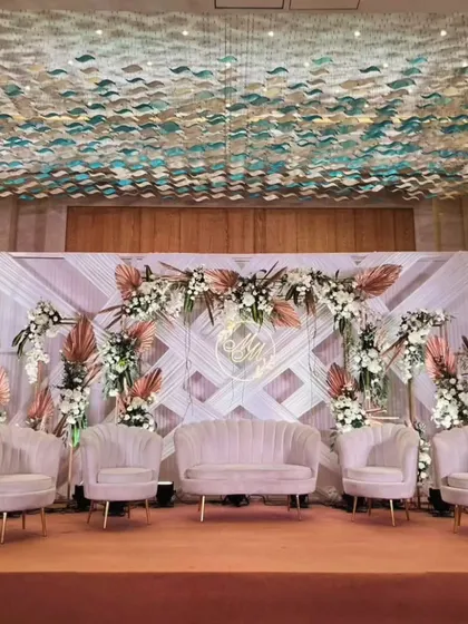 A sophisticated and modern reception stage design. The backdrop features a geometric pattern with pampas grass and pastel florals, complemented by plush pink seating and a custom neon initial sign.