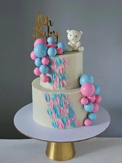 A classic baby shower or gender reveal cake in pink and blue. This two-tiered design features a cascade of balloons, a textured buttercream effect, and a sweet teddy bear topper.