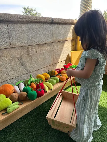 A child carries a wooden basket to do her "shopping" at our pretend vegetable market, engaging in imaginative role-play.