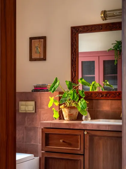 This bathroom in the Calcutta home features a warm, earthy palette. The terracotta-colored tiles, carved wooden mirror, and a potted plant create a space that feels grounded, natural, and calming.