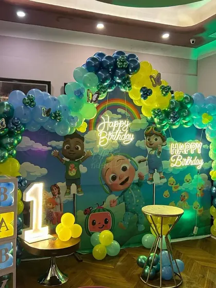 A Cocomelon first birthday decoration with a large backdrop and balloon arch. The setup includes a 'BABY' block prop and a marquee number '1'.