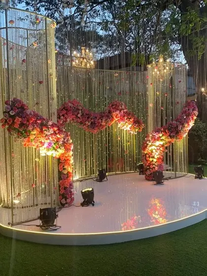 A unique and modern stage backdrop featuring a wavy structure adorned with crystals and a flowing arrangement of pink flowers. The spotlights from below highlight the texture and shape of the design.