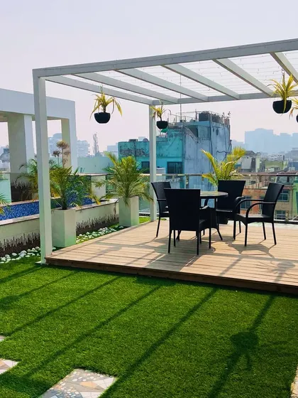 A rooftop escape in Dwarka. This design uses a clean, white metal pergola to create a shaded dining area. The space is defined by a wooden deck, artificial turf, and planters with palms, making it a perfect spot for enjoying the city view.