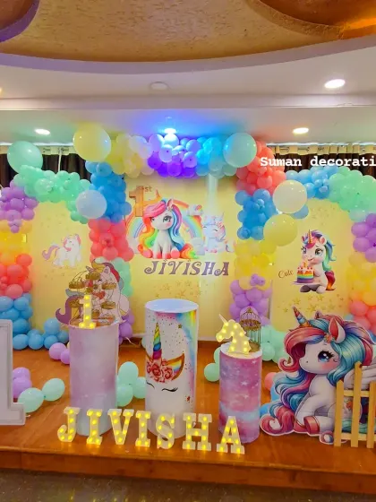 A magical unicorn-themed first birthday party. I used a pastel rainbow balloon arch, unicorn character cutouts, themed plinths, and the name 'Jivisha' in marquee letters.