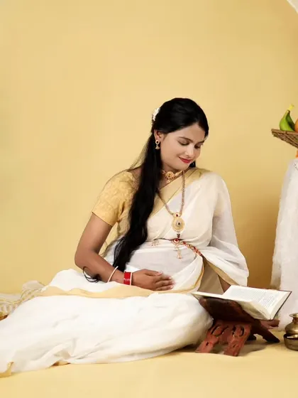A serene solo portrait from a South Indian themed maternity shoot. The mother-to-be is reading a book, surrounded by traditional elements like marigold flowers and a peacock backdrop.