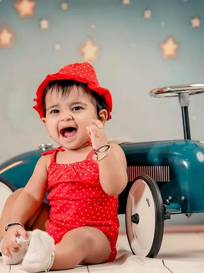 A happy little girl in a red outfit, sitting next to her vintage toy car. The balloon backdrop adds a touch of whimsy.
