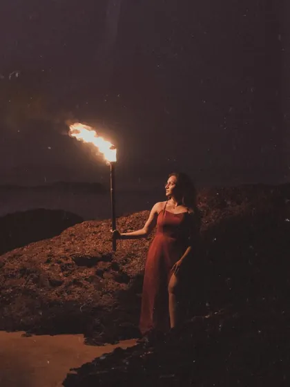 A full-length portrait of the bride-to-be in a red dress, standing by a rock and holding a flaming torch. The warm light of the flame illuminates her against the dark beach, creating a bold and beautiful image.