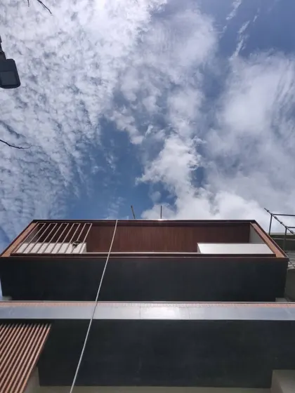 A low-angle shot of a residential apartment building under construction, with the sky as a dramatic backdrop. The image highlights the clean lines of the projecting wooden balcony against the dark stone cladding.