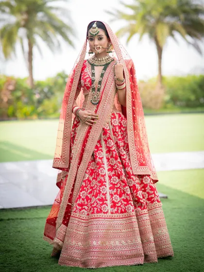 A full-length portrait of a bride looking absolutely majestic. The soft glam makeup with defined eyes stands out beautifully against her ornate red and gold lehenga, creating a truly royal vision.