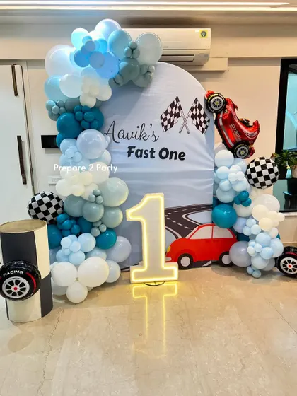 A "Fast One" car race theme for a first birthday. This setup includes a custom road-themed backdrop, a light-up marquee number, and a balloon garland with checkered flag details and a race car foil balloon.