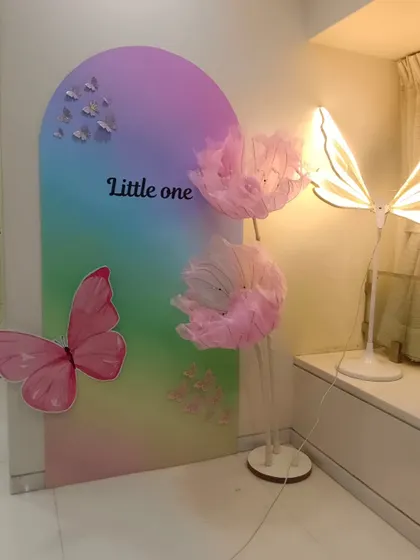 A modern art-style backdrop for a 'Little One' celebration, featuring a rainbow gradient panel, a large pink butterfly, and delicate light-up props.