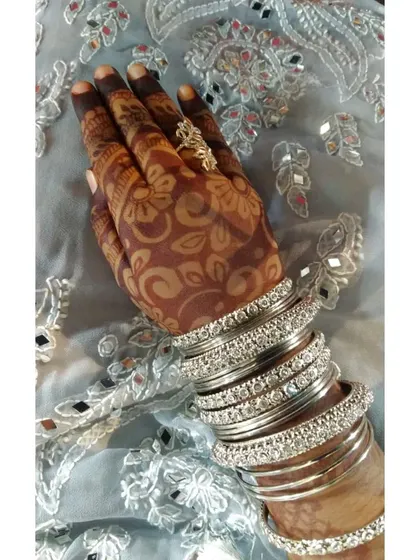 A close-up of a mature henna stain on the back of the hand, paired with sparkling silver bangles. The floral pattern is clear and the color is a deep reddish-brown, perfect for any celebration.