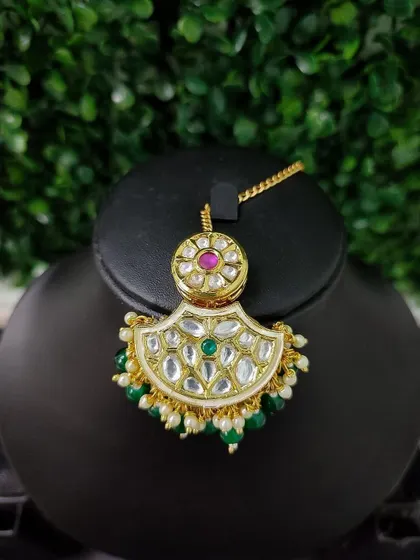 A simple yet beautiful crescent-shaped maang tikka with kundan work. The piece is accented with a central ruby, a green stone, and pearl drops.