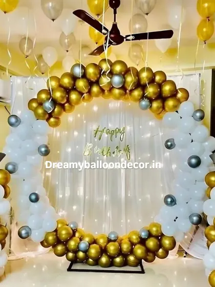 An elegant gold and white themed birthday backdrop. This home setup features a full balloon ring with chrome gold and white balloons, star-shaped foil balloons, and a sheer curtain with fairy lights.