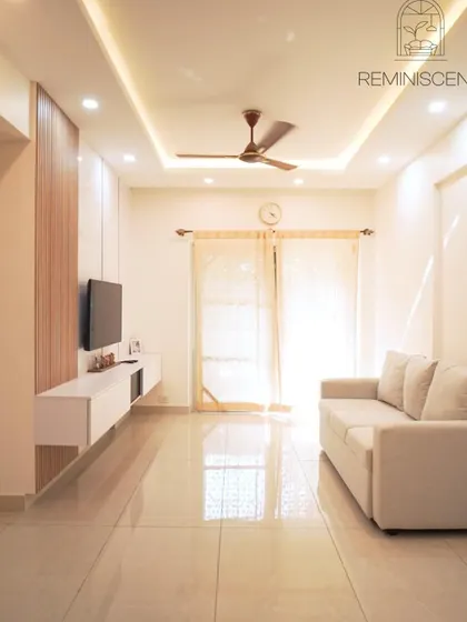 A bright, minimalist living room that feels spacious and airy. The simple white sofa, floating TV unit, and clean false ceiling design with recessed lighting create a serene and uncluttered environment.