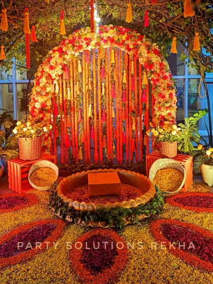 A stunning Haldi setup featuring a floral arch and a vibrant rangoli made of flower petals. The traditional brass urli at the center is the focal point for the ceremony.