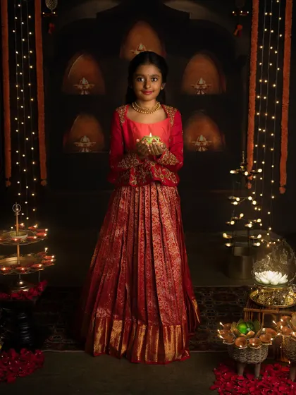 A young girl in a stunning red and gold lehenga holds a diya, her face lit with the joy of Diwali.