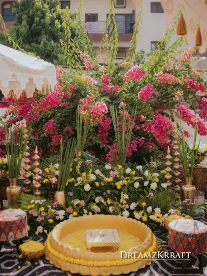 The Haldi seating area, set against a backdrop of vibrant pink bougainvillea and lush florals. The traditional brass 'urli' is ready for the ceremony.