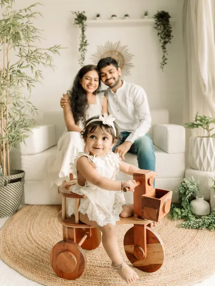 A one-year-old takes center stage on a wooden tricycle, with her proud parents watching from the background.