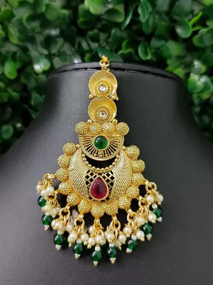 An antique-finish maang tikka with a crescent design. The red and green stones combined with pearl and green bead drops give it a vintage charm.